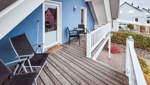 Eine Holzterrasse mit Gartenmöbeln, eine Glastür, die zu einem Raum führt, und ein weißes Geländer.