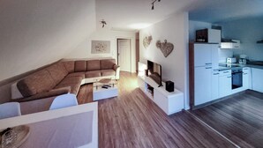 Ein modernes Wohnzimmer mit einem Ecksofa, einem Flachbildfernseher und einer minimalistischen Küche mit weißen Schränken und einem eingebauten Backofen.