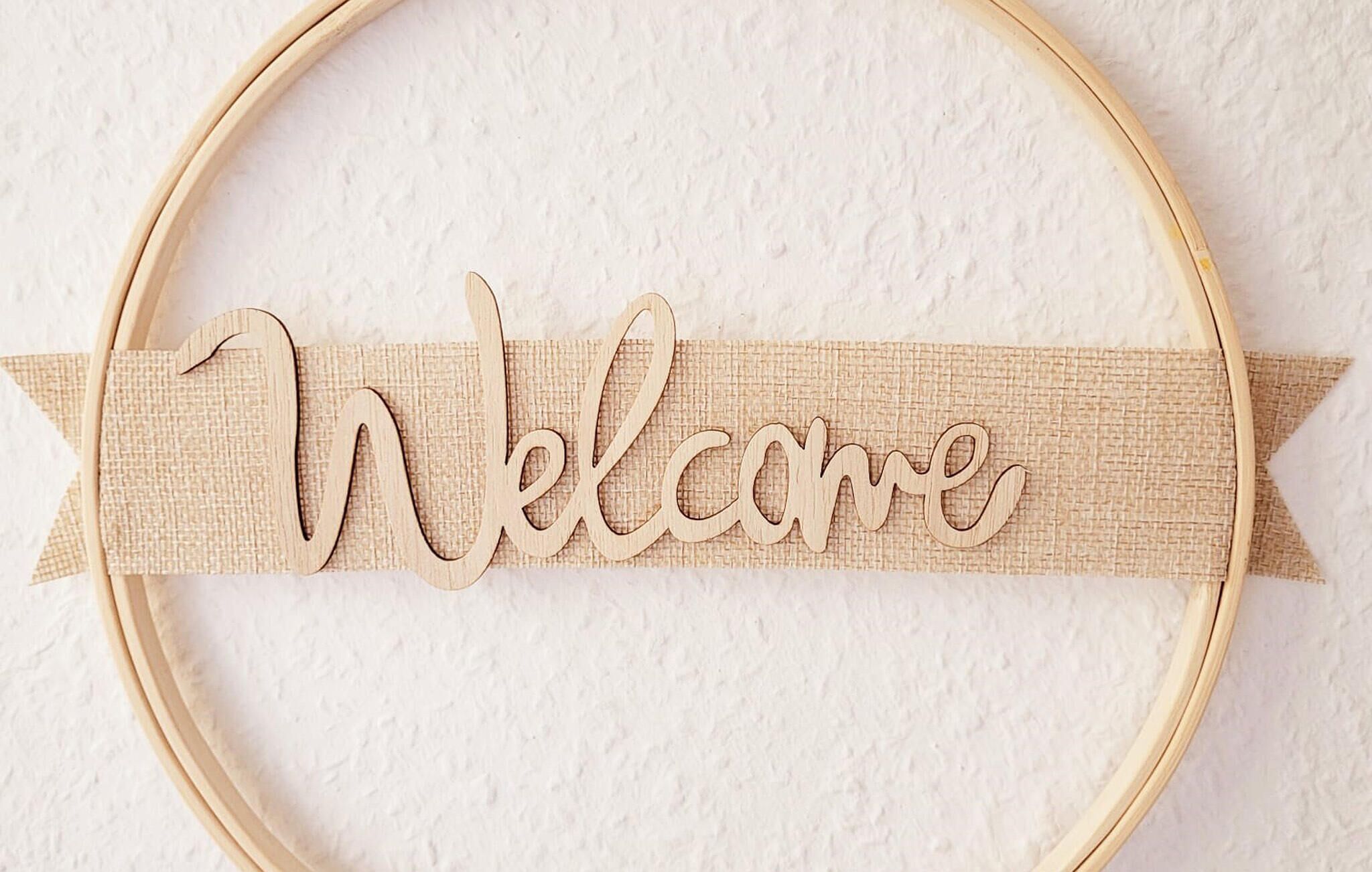 A wooden sign with the word "Welcome" on a burlap ribbon, placed inside a wooden frame.