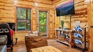 L’intérieur d’un chalet accueillant, avec des murs en bois, un téléviseur fixé au mur, une table de jeux et un coin salon comprenant un canapé et des fauteuils.