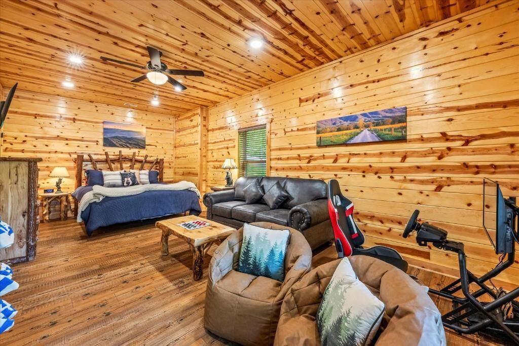 A log cabin interior with a bed, sofa, and armchair, wooden walls, and ceiling fan.