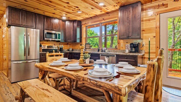 Une cuisine rustique aménagée dans une cabane en bois, avec une table à manger pour six personnes, des appareils en acier inoxydable et une grande fenêtre donnant sur un paysage verdoyant.