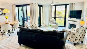 A modern living room with a black sofa, a dining table with chairs, a TV on a fireplace mantel, and large windows with curtains. - Beautiful 2 bedroom, 2 bath Destin, Florida Condo! (Destin)