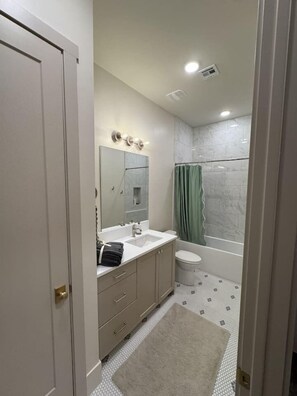 A bathroom with a white sink, mirror, and toilet, a shower with a green curtain, and a patterned floor.