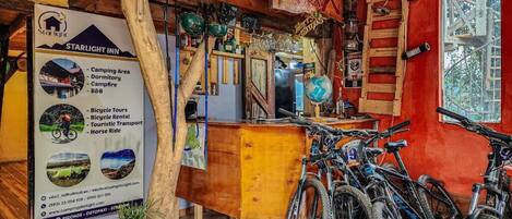 A rustic inn with a wooden interior, bicycles, and a signboard detailing amenities like camping, dormitory, and bicycle tours.