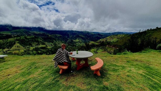 A person sitting at a round table with a view of rolling hills and a cloudy sky. - Starlight Mountain Lodge (Sigchos)