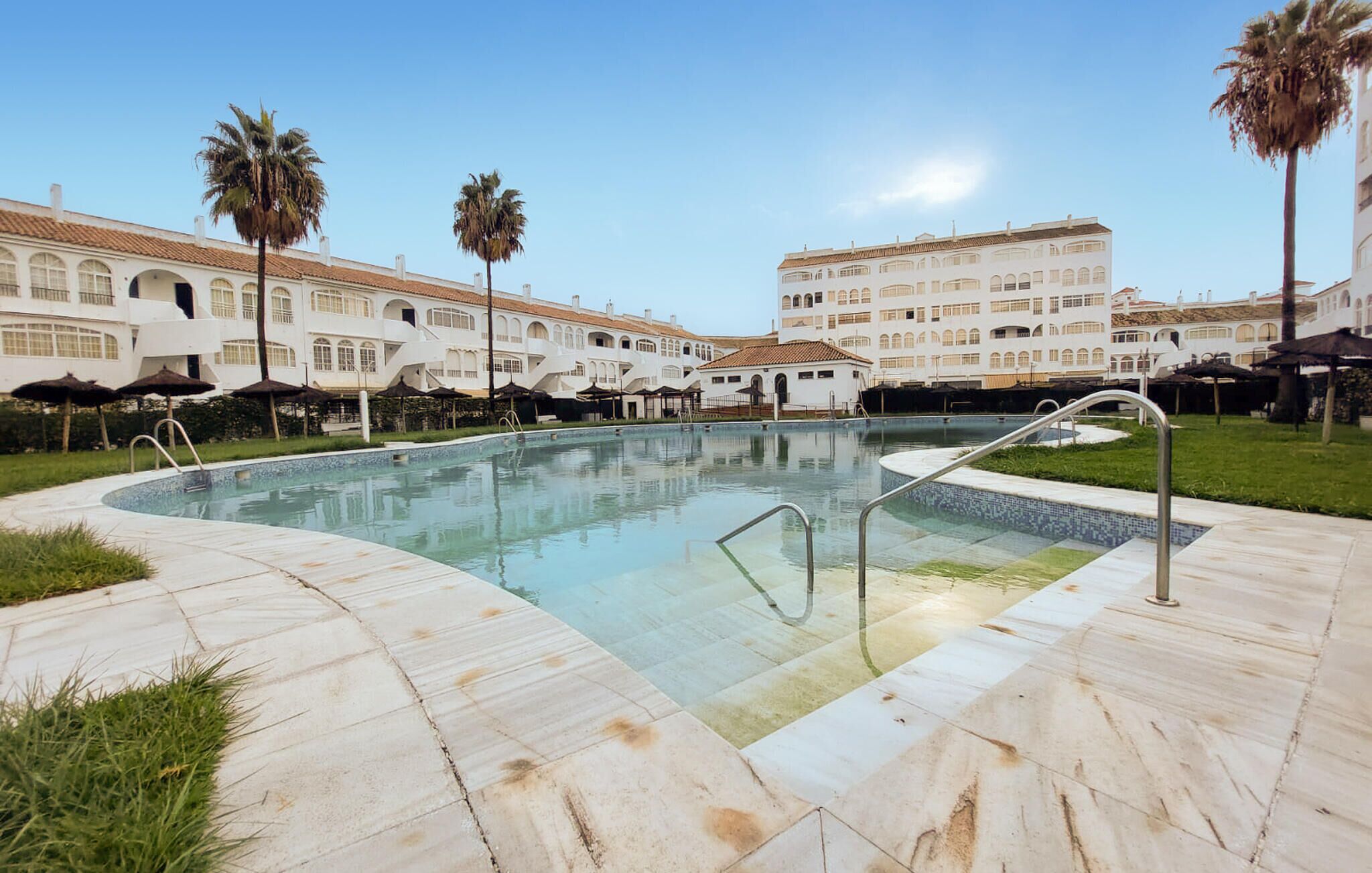 A swimming pool with a tiled deck, surrounded by palm trees and white buildings with arched windows.