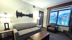 A hotel room with a sofa, armchair, coffee table, lamp, and a large window with a mountain view. - Modern condo at Falcon Crest Lodge with extra amenities (Canmore)
