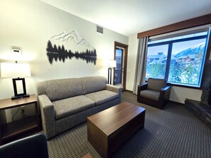 Ein Hotelzimmer mit einer Couch, einem Sessel, einem Couchtisch, einer Lampe und einem großen Fenster mit Blick auf die Berge.