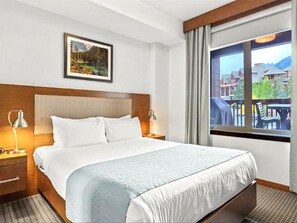 A hotel room with a large bed, a headboard, a nightstand with a lamp, a window with a view of buildings and mountains, and a framed picture on the wall. - Modern condo at Falcon Crest Lodge with extra amenities (Canmore)