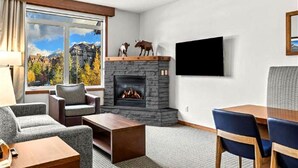 A modern living room with a stone fireplace, a television mounted on the wall, and a view of mountains through the window. - Modern condo at Falcon Crest Lodge with extra amenities (Canmore)