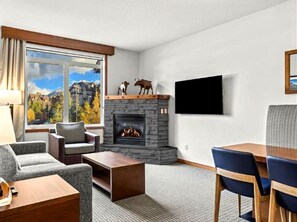 Ein modernes Wohnzimmer mit Kamin aus Stein, einem an der Wand befestigten Fernseher und Blick auf Berge durch das Fenster.