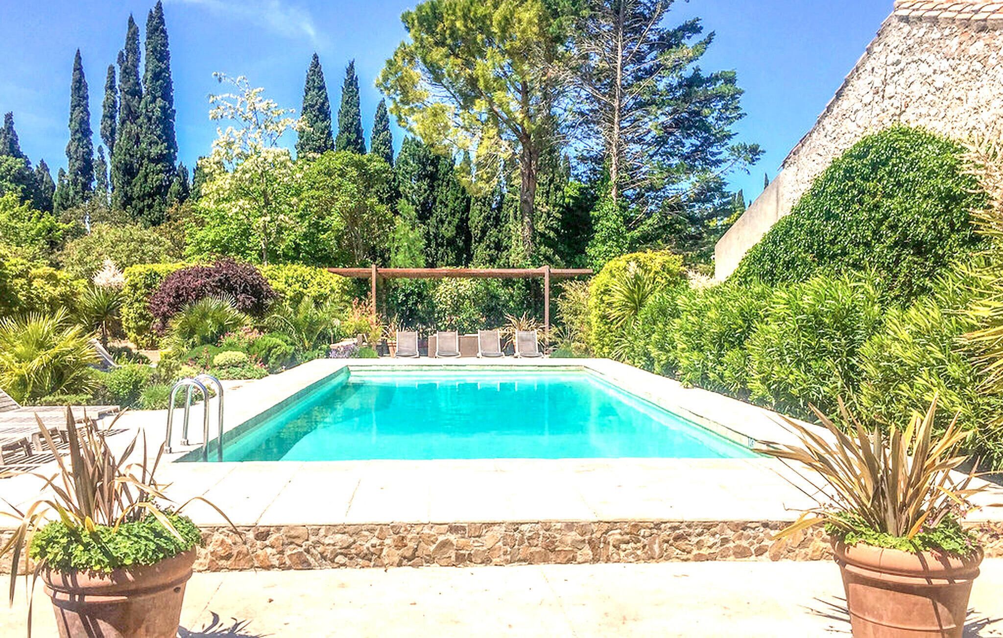 Une piscine rectangulaire agrémentée d’une pergola et de chaises, entourée d’une végétation luxuriante et de grands arbres.