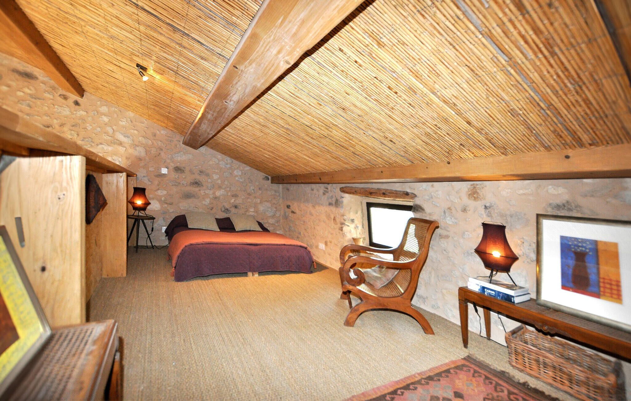 Ein gemütliches Schlafzimmer im Dachgeschoss mit Steinwand, Holzbalkendecke, einem Bett, einem Sessel, einem Tisch mit Lampe und einem gerahmten Bild.