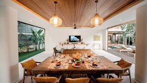 A dining area with a wooden table set for a meal, a television in the background, and a view of a garden through large windows. - Villa Seminyak 4BR w/pool close to Petitenget (Badung Regency)