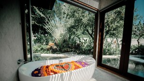 A modern bathroom with a freestanding bathtub filled with colorful flowers, large windows, and a view of lush greenery. - Villa Seminyak 4BR w/pool close to Petitenget (Badung Regency)