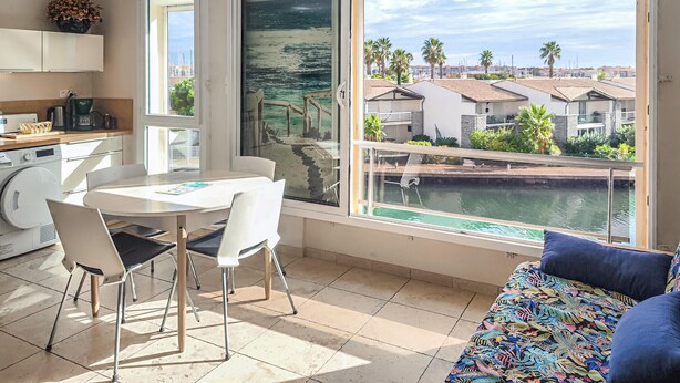 A modern kitchen with a coffee machine, a dining area with a table and chairs, and a large window with a view of a marina and buildings. - Cozy apartment in Agde with house sea view (Agde)
