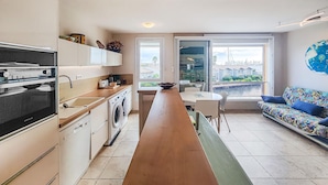 A compact kitchen with a long wooden countertop, a washing machine, and a dining area with a bench and table. - Cozy apartment in Agde with house sea view (Agde)