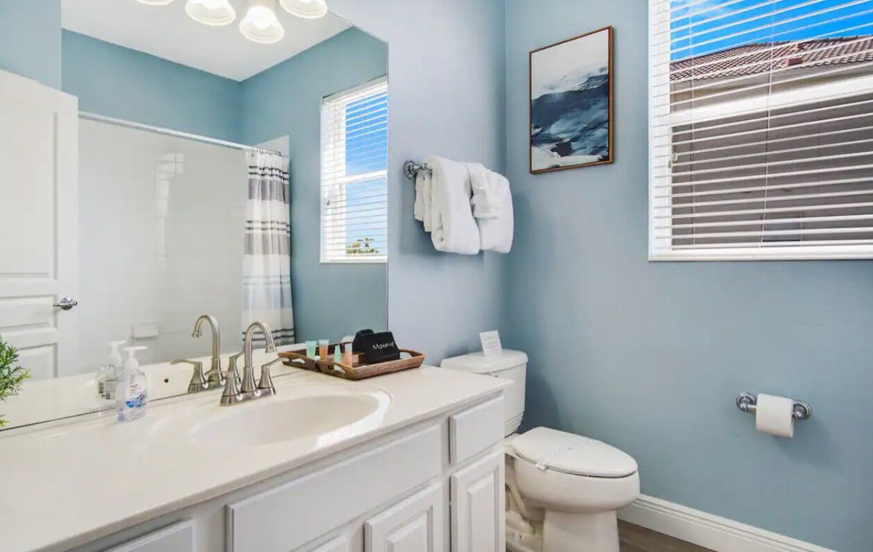 Une salle de bain avec un lavabo blanc, deux robinets, un miroir, des toilettes et une fenêtre avec des stores.