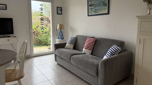 A living room with a grey sofa, a television, a dining table, and chairs. There is a door leading to a garden.