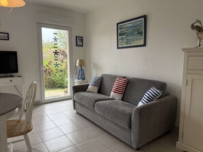 Una sala de estar con un sofá gris, un televisor, una mesa de comedor y sillas. Hay una puerta que da a un jardín.