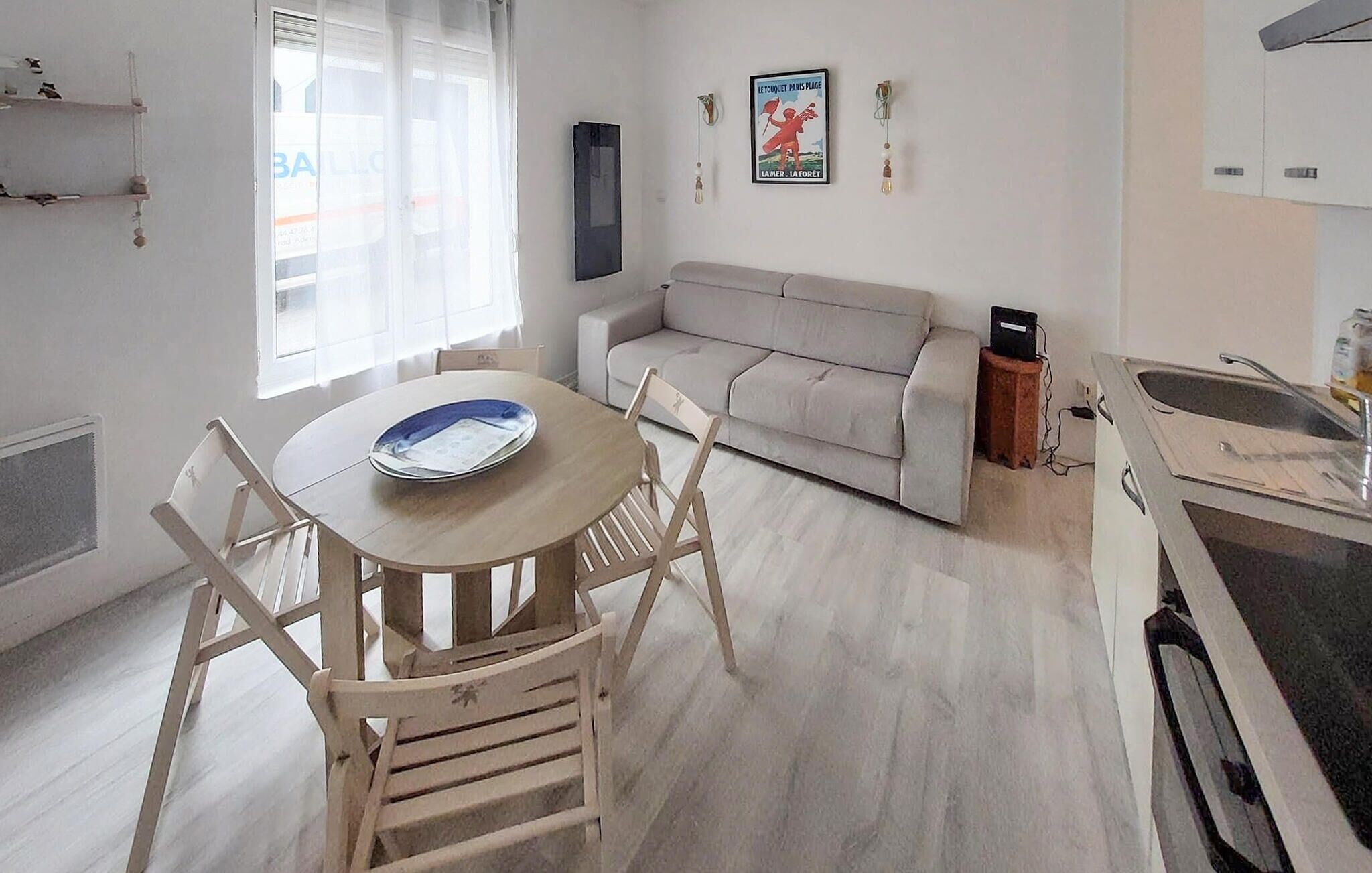 Ett modernt vardagsrum med en grå soffa, ett matbord i trä med stolar, en köksdel med diskho och spis samt en affisch på väggen.
