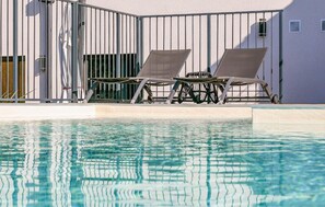 Ett poolområde med två solstolar och ett bord, omgivet av en vit byggnad med ett staket av metall.