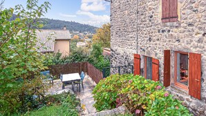 Un edificio in pietra con persiane di legno, una piccola area esterna con un tavolo e sedie, e vegetazione lussureggiante.