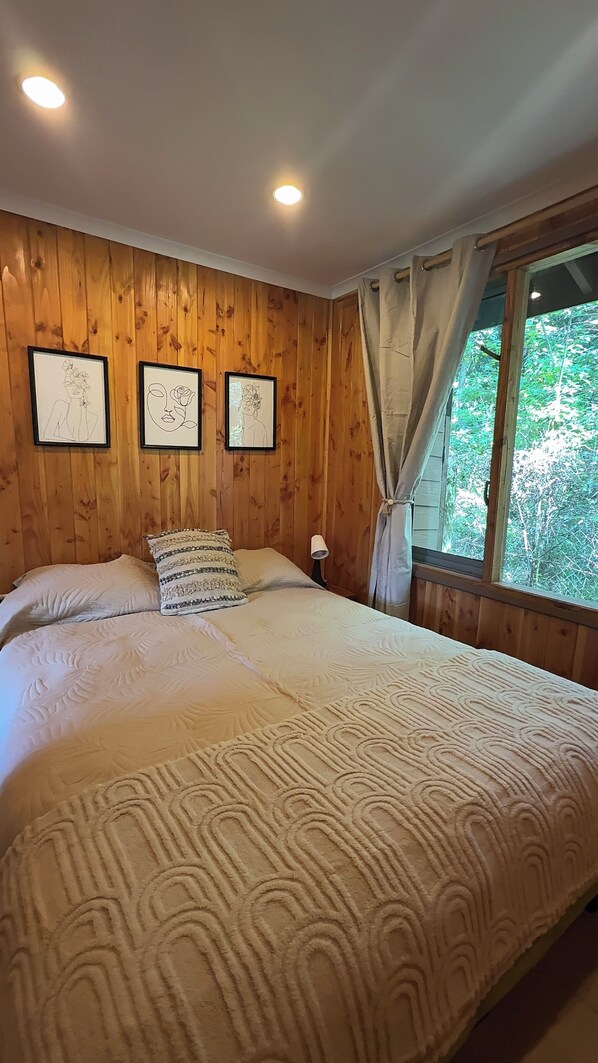 A wooden-paneled room with a large bed, three framed artworks on the wall, and a window with curtains. - Casa Tuwa Cabañas en pucón (Pucón)