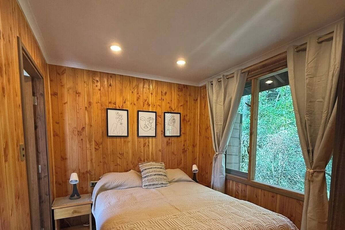 A wooden-paneled bedroom with a bed, a nightstand, and three framed artworks on the wall.