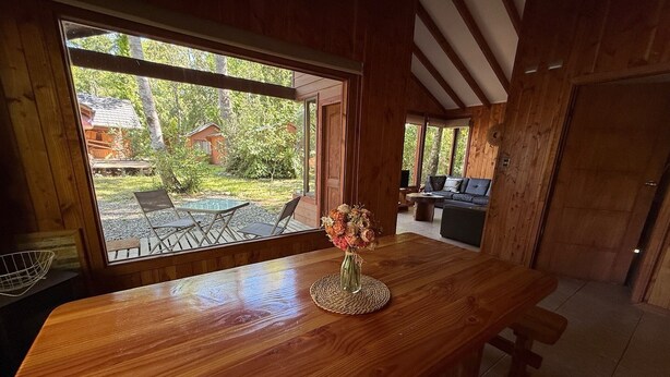 A wooden interior with a large window, a wooden table with a vase, and a view of an outdoor seating area. - Casa Tuwa Cabañas en pucón (Pucón)