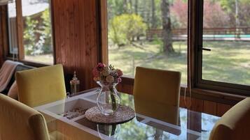 Un comedor con mesa de vidrio, sillas amarillas, un jarrón con flores y vistas a árboles a través de amplias ventanas.