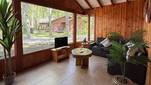 A spacious living room with wooden walls and large windows, a black sofa, a coffee table, and a TV on a wooden stand. - Casa Tuwa Cabañas en pucón (Pucón)