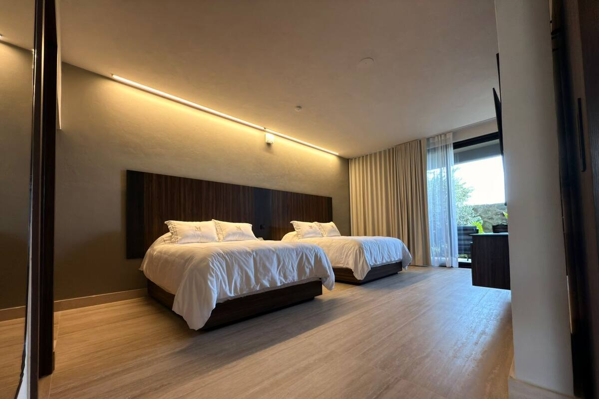 Une chambre d’hôtel moderne avec deux lits, un plancher en bois, une grande fenêtre avec des rideaux et une vue sur la verdure.