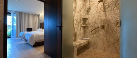 Una habitación de hotel moderna con una pared de mármol, suelo de madera, una cama grande y un baño con ducha de mármol.