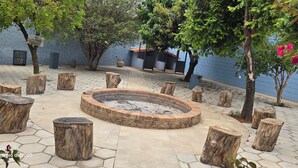 A circular fire pit surrounded by wooden stumps and a paved patio area with trees and a blue wall in the background. - BomjaMinas property, swimming pool, bed linens and towels, 17 km from Inhotim. (Mário Campos)