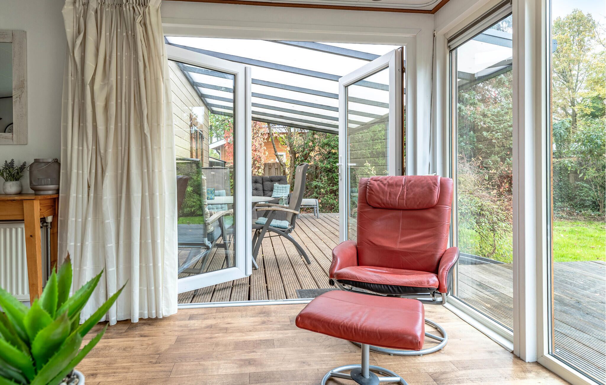 A conservatory with a red leather chair and a glass table, wooden flooring, and a view of a garden.