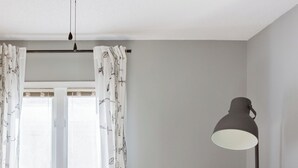 Um quarto com janela, cortinas brancas, uma luminária, um sofá e uma placa de madeira.
