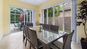 A dining area with a glass table and wicker chairs, large windows, and a ceiling fan. - 🏖️ 4BR Beach House w/ Heated Spa and Pool (Naples)
