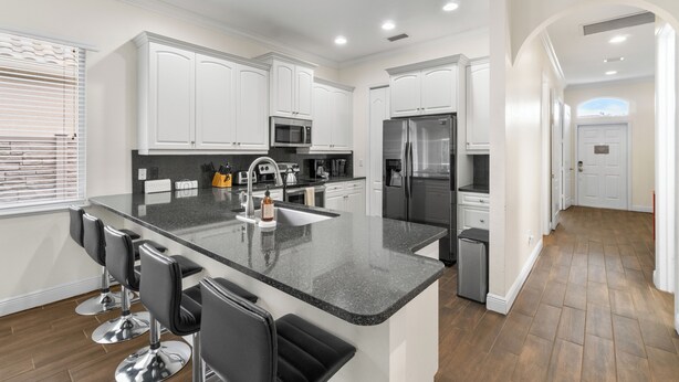 A modern kitchen with white cabinets, a granite countertop, and stainless steel appliances. There are bar stools and a refrigerator. - 🏖️ 4BR Beach House w/ Heated Spa and Pool (Naples)