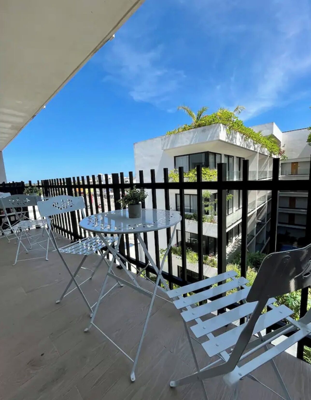 En balkon med metalstole og et rundt bord, med udsigt over en moderne bygning med balkoner og grønt.