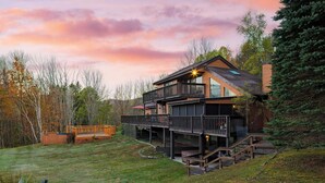 Una casa a due piani in legno con veranda e area esterna coperta, immersa tra alberi e un prato.