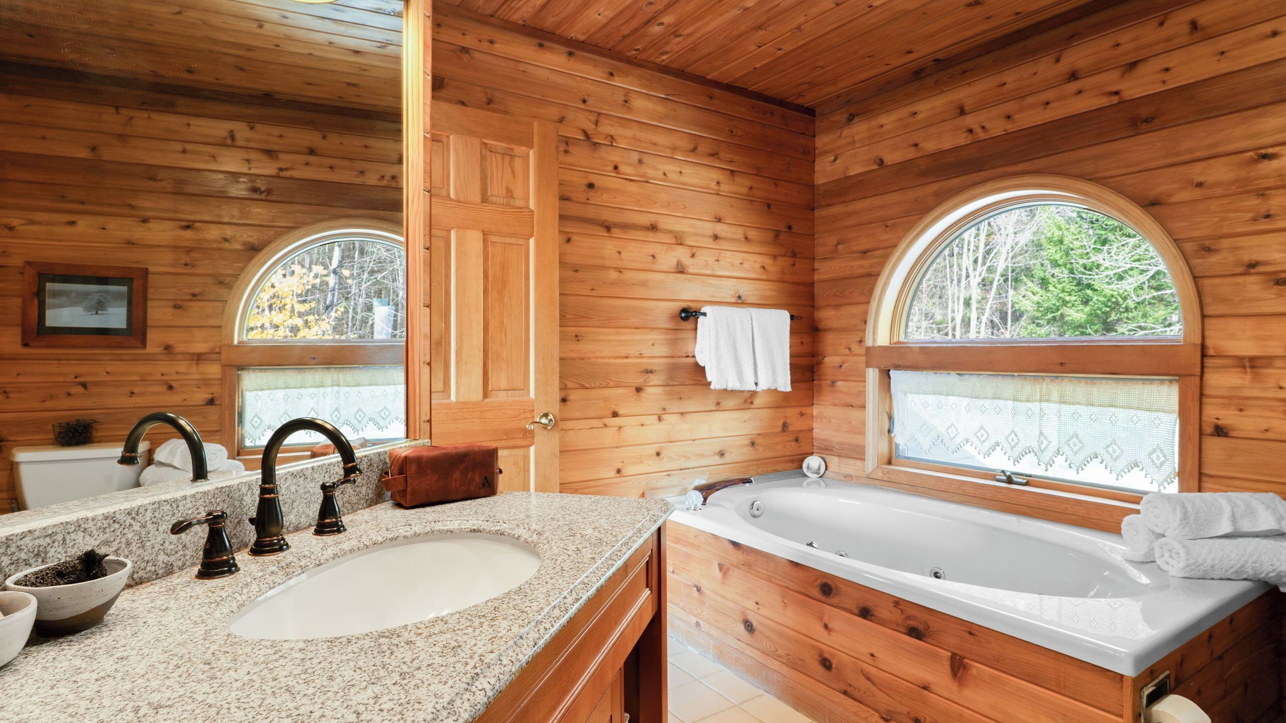 A wooden cabin bathroom with a sink, two faucets, a bathtub, and two windows.