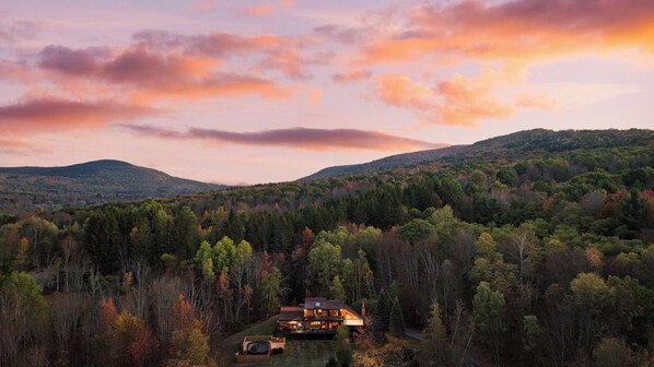 Una casa grande rodeada de un bosque denso, con vistas a colinas ondulantes al fondo.