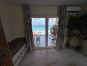 Een kamer met een schuifglasdeur die leidt naar een balkon met uitzicht op het strand en de oceaan. Op het balkon staat een rieten stoel en een klein tafeltje.