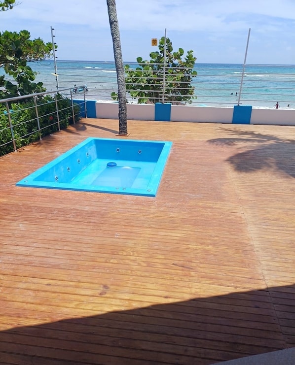 Een houten terras met een blauw zwembad, omringd door een metalen hek en torenhoge palmbomen, met uitzicht op de oceaan.