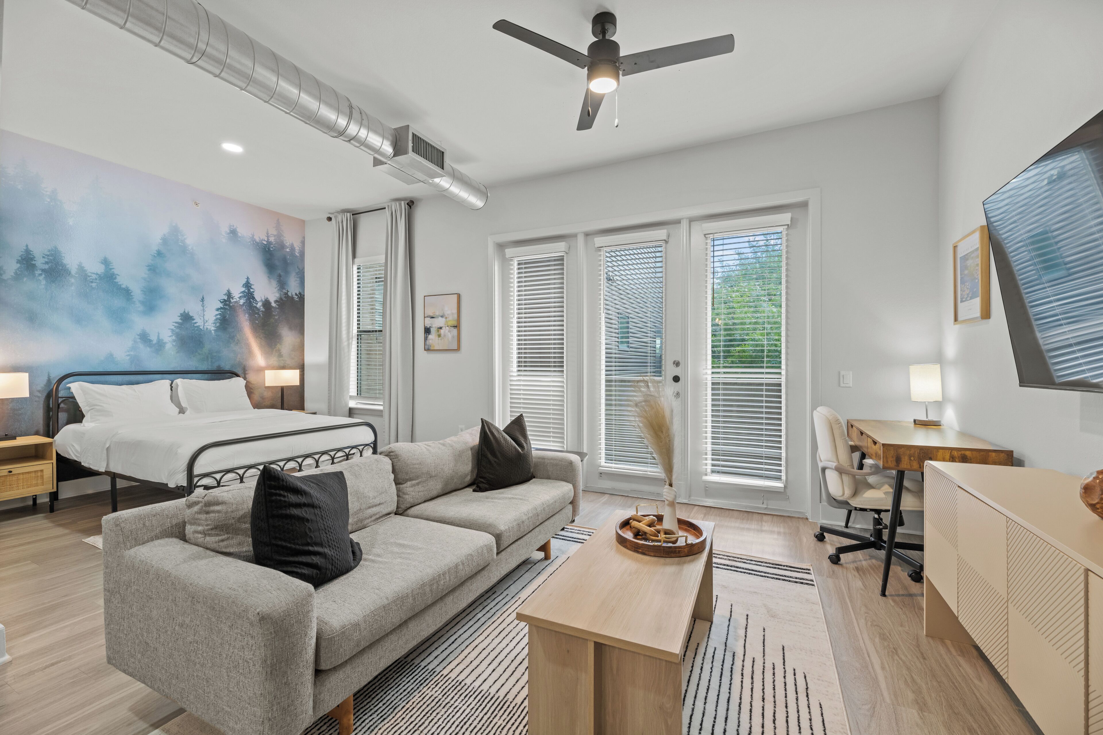 Une chambre moderne avec un lit, un canapé gris, un bureau en bois et une grande fenêtre avec des stores.