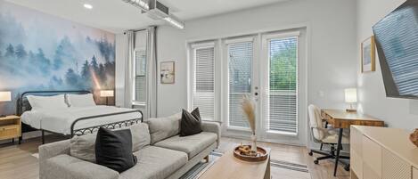 Une chambre moderne avec un lit, un canapé gris, un bureau en bois et une grande fenêtre avec des stores.