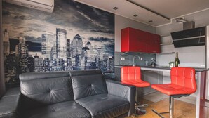 Un salón moderno con un sofá de cuero negro, dos taburetes rojos, un mural con vista a la ciudad y una cocina con armarios rojos y una campana extractora de acero inoxidable.