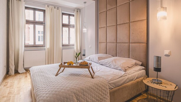 Un dormitorio con cama, una mesita de noche con lámpara, una ventana con cortinas y una bandeja con comida sobre la cama.